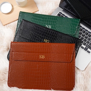 May include: Three personalised laptop sleeves in green, black, and brown, with a crocodile skin texture. Each sleeve has initials: "NB", "HG", and "SB". A laptop and a lit candle are also visible.