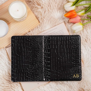 May include: Black leather notebook with a crocodile skin texture, personalized with gold initials "AB". The notebook is open, revealing blank pages. The background includes a wooden tray, candles, and tulips.
