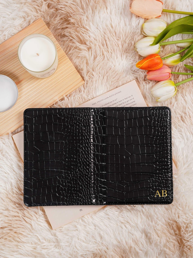May include: Black leather notebook with a crocodile skin texture, personalized with gold initials "AB". The notebook is open, revealing blank pages. The background includes a wooden tray, candles, and tulips.