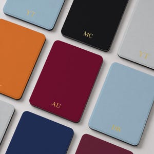 May include: A collection of rectangular passport holders in various solid colours including light blue, black, grey, orange, dark red, and navy blue. Each holder features gold-embossed initials such as YT, MC, AU, and DB centred on the front cover.