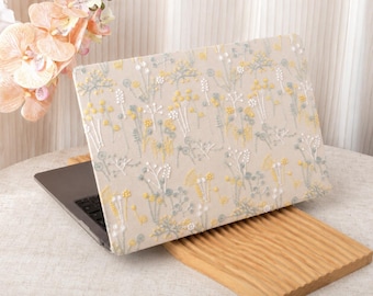 May include: A laptop computer featuring a protective cover with a cream-colored fabric base and an embroidered pattern of small yellow and light blue wildflowers. The laptop is positioned on a light-colored wooden stand with a ribbed surface texture.