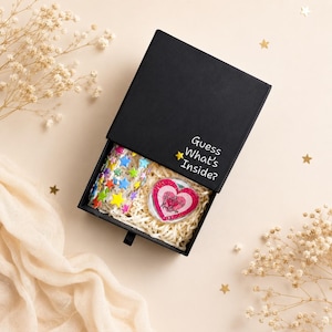 May include: A black sliding gift box with the text Guess What's Inside? printed in white, featuring a small yellow star accent. Inside, the box contains a heart-shaped item with pink and red layers and a sheet of colorful star stickers.