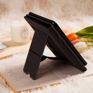 May include: A dark brown tablet case with a stand, featuring white stitching. The case is propped open on a book with text, set against a soft, white, fluffy background. A candle and flowers are also visible.