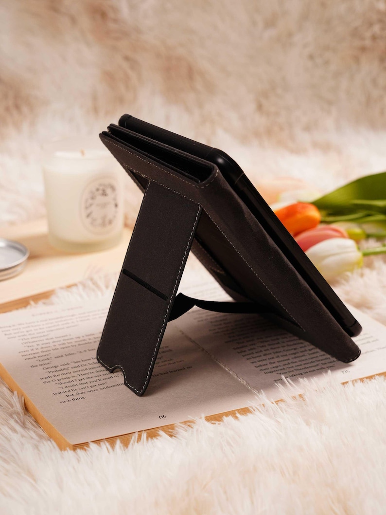 May include: A dark brown tablet case with a stand, featuring white stitching. The case is propped open on a book with text, set against a soft, white, fluffy background. A candle and flowers are also visible.