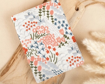 Pode incluir: Uma carteira de tecido branco com um design de bordado floral em tons de rosa, vermelho e azul. A carteira tem uma superfície texturizada e um brilho sutil.