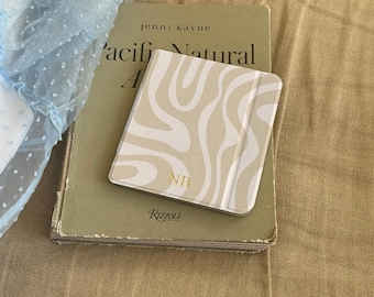 May include: A square tablet case with a beige and white wavy line pattern and the gold embossed letters NB, resting on a hardcover book titled Pacific Natural by Jenni Kayne published by Rizzoli, next to a light blue sheer fabric with white dots.