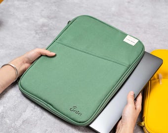 May include: A sage green laptop sleeve with a zip closure and a small front pocket. The sleeve has the word "Scase" printed on the lower right corner. A laptop is partially inserted into the sleeve. A yellow laptop sleeve is visible in the background.