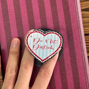 Op de afbeelding: Een hartvormige telefoonhouder met de tekst "Do Not Disturb" in rood script. De houder heeft een witte geschulpte rand, een lichtblauwe en wit gestreepte achtergrond en een zwarte basis. De houder staat op een gestreepte achtergrond.