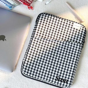 May include: A black and white gingham laptop sleeve with the name "Sasa" printed on the front. Other gingham cases in red and blue are visible. A silver laptop, charger, and stylus are also in the image.