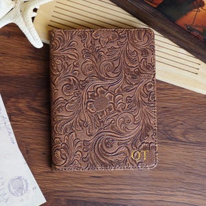 Peut inclure: Un journal en cuir marron avec un motif floral en relief. Le journal porte un monogramme initial doré. Il est posé sur une surface en bois, avec une étoile de mer et une œuvre encadrée en arrière-plan.