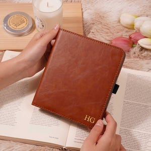 May include: A brown leather journal with the gold initials "HG" is held open over a book. A candle in a glass jar and a small tin are in the background. The journal has a black elastic band.