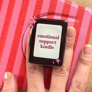 Pode incluir: Um anel retangular preto com as palavras "emotional support kindle" em fonte roxa. O anel está em um dedo, com um fundo listrado rosa e vermelho.