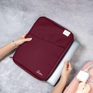 May include: A claret laptop sleeve with a front pocket and a zip closure. The sleeve has the word "Sasa" embroidered in white script. A small white tag with text is on the upper right corner. A silver laptop is partially inside the sleeve.