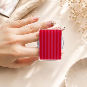 May include: A rectangular phone grip featuring a vibrant pattern of alternating vertical stripes in shades of bright red and deep magenta. The accessory has a smooth, glossy finish and is mounted on a circular base, designed to attach to a mobile device.