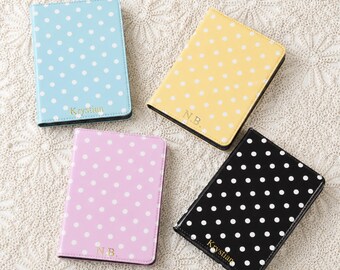 May include: Four rectangular passport holders featuring a polka dot pattern in light blue, yellow, light purple, and black. Each holder displays a gold-coloured name or initials, specifically Krystian on the blue and black covers, and N.B. on the yellow and purple covers.