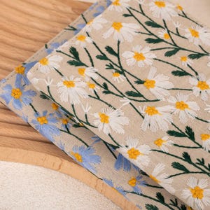 Pode incluir: Duas amostras de tecido com padr&otilde;es florais bordados. A amostra superior &eacute; bege com margaridas brancas e caules verdes. A amostra inferior &eacute; azul com flores azuis claras.