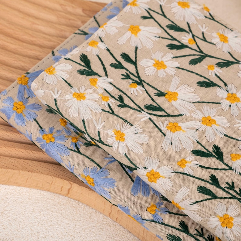 Pode incluir: Duas amostras de tecido com padr&otilde;es florais bordados. A amostra superior &eacute; bege com margaridas brancas e caules verdes. A amostra inferior &eacute; azul com flores azuis claras.