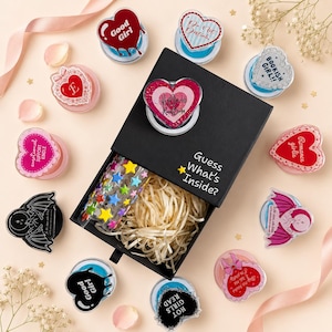 May include: A black gift box with the text Guess What's Inside? containing shredded paper and a sheet of star stickers, surrounded by various heart-shaped acrylic pins featuring designs like Good Girl, Bookish Girl, and Romance Girlie.
