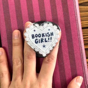 May include: A heart-shaped phone grip with the text "BOOKISH GIRL!!" in navy blue, surrounded by a white lace-like border and small stars. The grip is held over a dark pink and burgundy striped surface.
