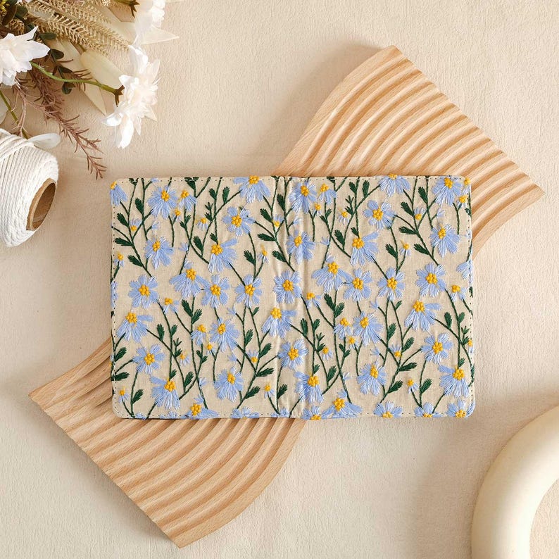 Pode incluir: Um pequeno caderno de cor creme com um design floral. O caderno &eacute; coberto com um padr&atilde;o floral azul claro e verde com pequenas flores brancas.