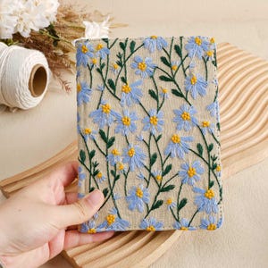 Pode incluir: Uma pequena bolsa de linho bege com fecho de correr, bordada com um padr&atilde;o floral de flores azuis e amarelas.