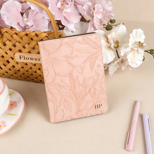 May include: A pink leather journal with a floral embossed design and the initials "BP" on the front cover. The journal is sitting on a light brown surface with a wicker basket and pink flowers in the background.