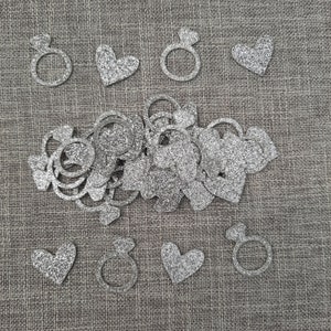 May include: Silver glitter confetti with heart and ring shapes. The confetti is scattered on a brown burlap background.