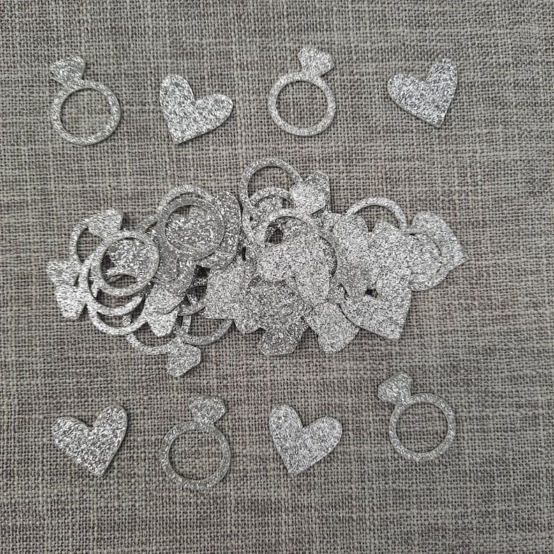 May include: Silver glitter confetti with heart and ring shapes. The confetti is scattered on a brown burlap background.