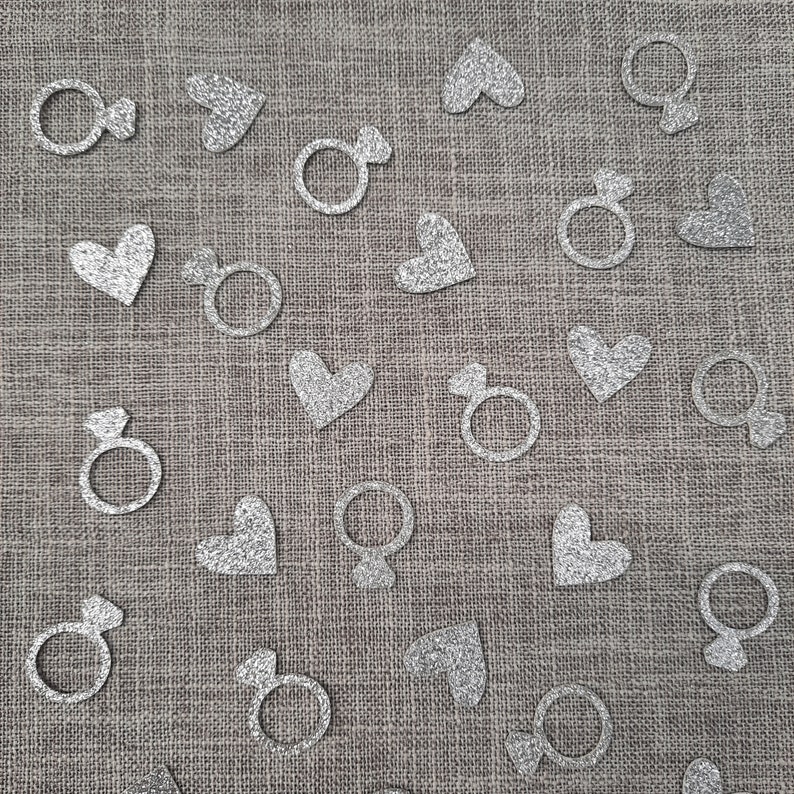 May include: Silver glitter confetti with heart, ring, and gender symbols on a gray burlap background.