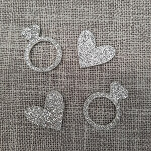 May include: Four silver glitter shapes on a brown burlap background. The shapes are a ring, a heart, a ring, and a heart.