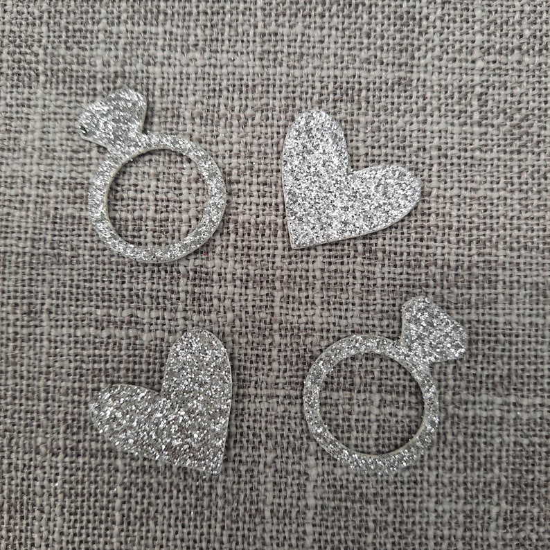 May include: Four silver glitter shapes on a brown burlap background. The shapes are a ring, a heart, a ring, and a heart.