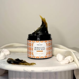 May include: A jar of Moroccan black soap with Argan oil. The jar has a black lid and a patterned label with the words "Moroccan Black Soap" and "With Argan". The soap is dark brown and has a thick, textured consistency. The jar contains 200 g of soap.