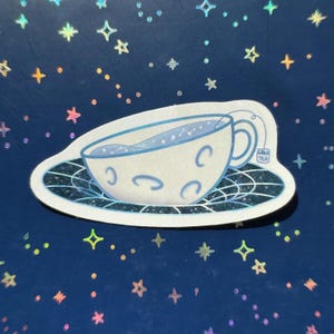 Puede incluir: Una pegatina con una taza de té blanca con líquido azul y diseños de media luna, sobre un platillo con un patrón en blanco y negro. La taza de té tiene una etiqueta que dice "Gravi Tea". El fondo es azul oscuro con diseños de estrellas y puntos.