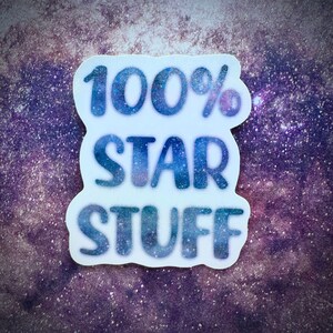 May include: A white sticker with the text "100% STAR STUFF" in blue with a glittery effect. The sticker has a unique shape and is set against a purple and white galaxy background.