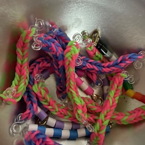 May include: A collection of colourful rubber band bracelets with different patterns and colours, including pink, blue, green, and white. The bracelets are made with a braided design and have clear plastic clasps.