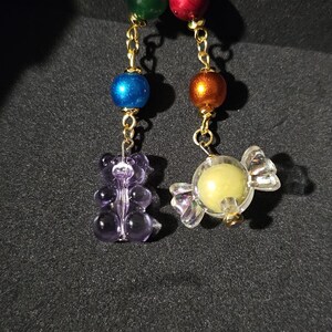 May include: A gold chain with colorful beads and charms, including a purple gummy bear and a clear candy. The charms are hanging from a gold clasp.