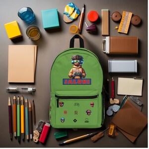 Pode incluir: Uma mochila verde com um bolso com z&iacute;per preto e uma al&ccedil;a superior. A mochila apresenta um gr&aacute;fico de um personagem segurando um controle de jogo, o nome "JALONI" e adesivos com tema de jogo. V&aacute;rios materiais escolares circundam a mochila.