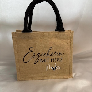 May include: A brown burlap tote bag with black handles. The bag has the text "Erzieherin mit Herz Melissa" printed on the front.