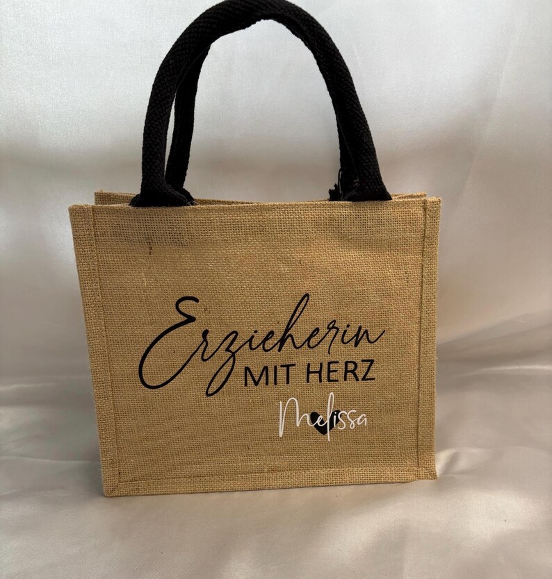May include: A brown burlap tote bag with black handles. The bag has the text "Erzieherin mit Herz Melissa" printed on the front.