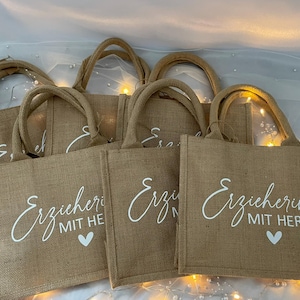 May include: Five brown burlap tote bags with white handles and white text that reads "Erzieherin mit Herz" and "Erzieherin mit He" on the front of the bags. The bags are arranged in a stack.