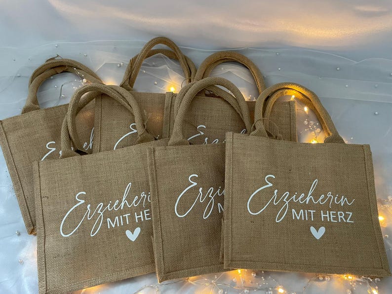 May include: Five brown burlap tote bags with white handles and white text that reads "Erzieherin mit Herz" and "Erzieherin mit He" on the front of the bags. The bags are arranged in a stack.