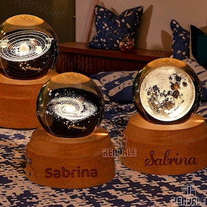 May include: Three glass globes with different celestial designs on wooden bases. The globes are lit from within, creating a warm glow. The bases are engraved with the name "Sabrina".