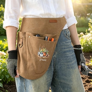 May include: A tan canvas garden tool belt featuring a large pocket with a printed graphic of a trowel, a hand rake, and a potted plant above the name Penny. The belt includes a metal D-ring on the side and multiple slots for holding small tools.