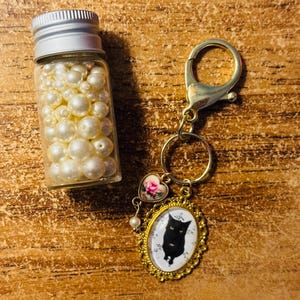 May include: A small glass bottle filled with white pearls sits next to a gold-colored keychain. The keychain has a charm with a black cat silhouette in an oval frame, and a heart-shaped charm with pink roses.