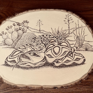 May include: A wood slice featuring a detailed pointillism illustration of a coiled rattlesnake in a desert landscape. The snake is black and white, with a detailed pattern. Cacti and desert plants are also depicted.