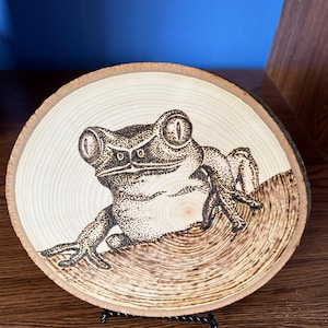 May include: A wood-burned art piece featuring a detailed frog design. The frog is depicted with intricate shading and sits on a wooden slice, showcasing the natural wood grain. The artwork is displayed on a black metal stand.