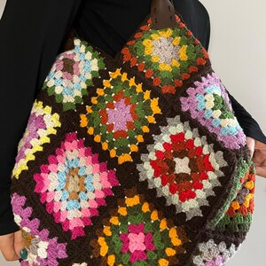Può includere: Una grande borsa tote all'uncinetto fatta a mano con un design patchwork di quadrati granny colorati. La borsa ha un colore di base marrone scuro con quadrati nelle tonalità di rosa, giallo, verde e blu. Ha manici in pelle marrone.