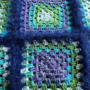 May include: A crocheted blanket with a blue border and squares in shades of green, purple, and blue.