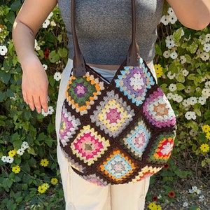 May include: A brown crocheted tote bag with colorful granny squares. The bag has leather straps and is made with a variety of colors, including pink, yellow, blue, green, and white.