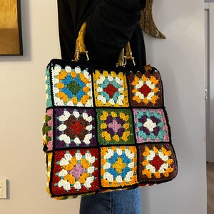 May include: A colorful crocheted tote bag with a brown wooden handle. The bag is made of squares in various colors, including red, orange, yellow, green, blue, and purple.